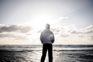 A man in a white jacket stands on the shore, facing the ocean at sunrise or sunset, with bright sunlight breaking through the clouds.